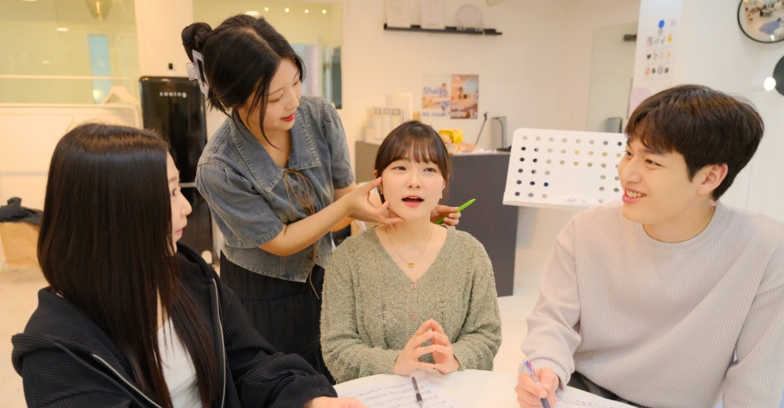 흰 라운지 원형 테이블에서 강사가 수강생의 턱을 짚어 주고 다른 수강생 둘이 악보를 들고 지켜보는 모습