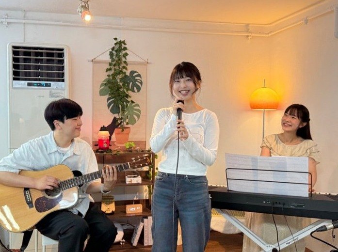 따뜻한 조명이 켜진 레슨실에서 통기타를 치는 남성과 마이크 앞에서 노래하는 여성, 건반 앞에 앉은 여성이 함께 연주하는 모습