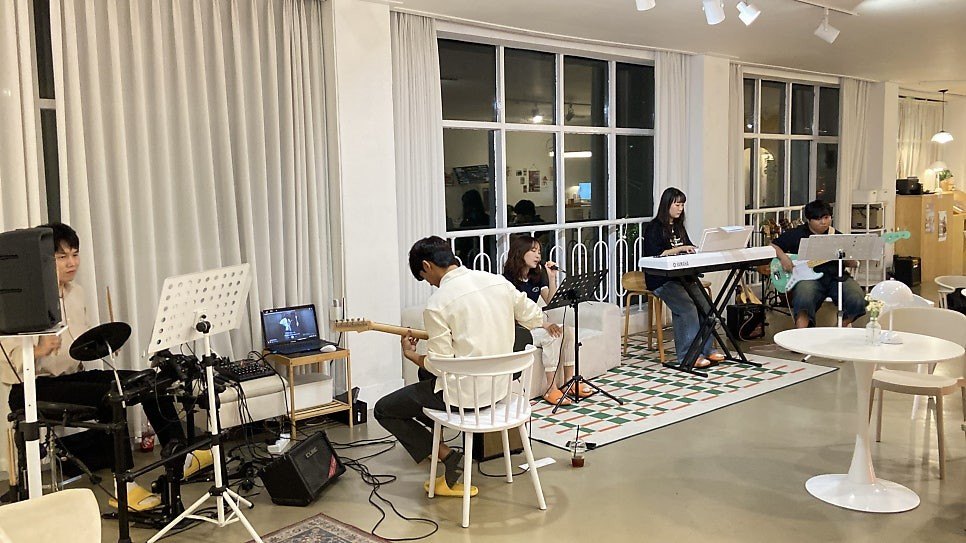 흰 커튼이 드리운 라운지에서 저녁에 드럼·기타·건반·베이스 연주자와 보컬이 함께 합주하는 모습, 체크무늬 러그와 보면대