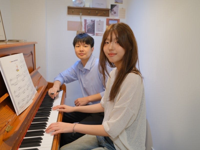 원목 업라이트 피아노 앞에 나란히 앉은 강사와 수강생, 보면대에 펼쳐진 악보와 벽에 붙은 엽서들
