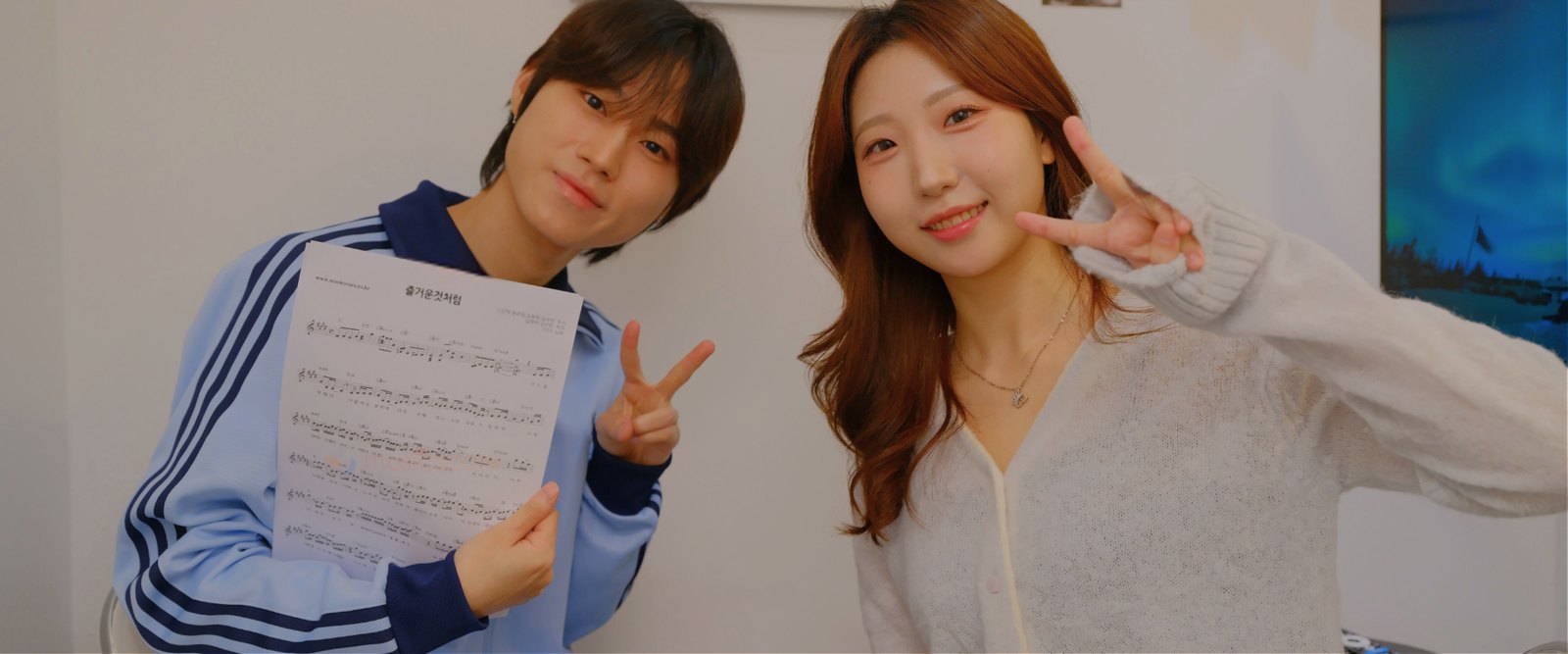 흰 벽 앞에서 악보를 든 학생과 강사가 나란히 서서 브이 포즈를 하며 웃는 모습