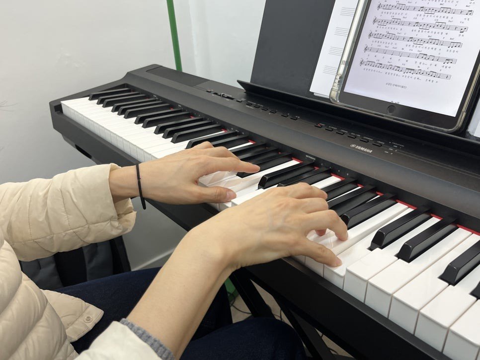 패딩 차림의 수강생이 태블릿에 띄운 악보를 보며 검은 디지털 피아노 건반에 두 손을 올린 모습