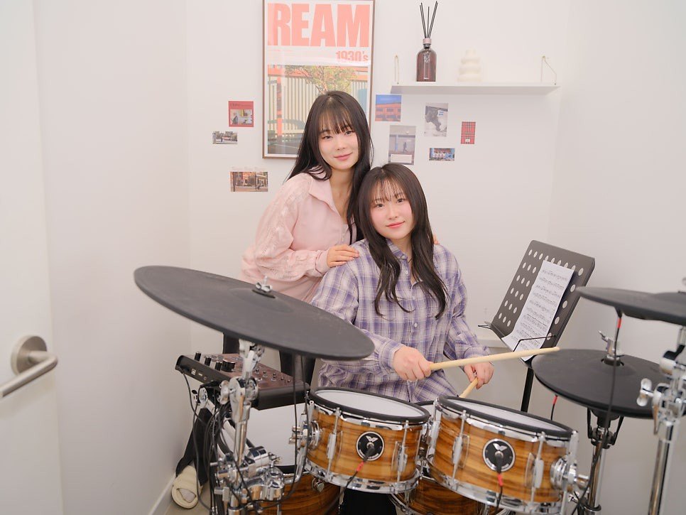 흰 벽의 작은 레슨실에서 체크 셔츠를 입은 수강생이 전자 드럼 앞에 앉아 스틱을 들고, 분홍 니트를 입은 사람이 뒤에서 어깨에 손을 얹고 있는 모습. 왼쪽에 문손잡이와 오른쪽에 악보 보면대가 보인다
