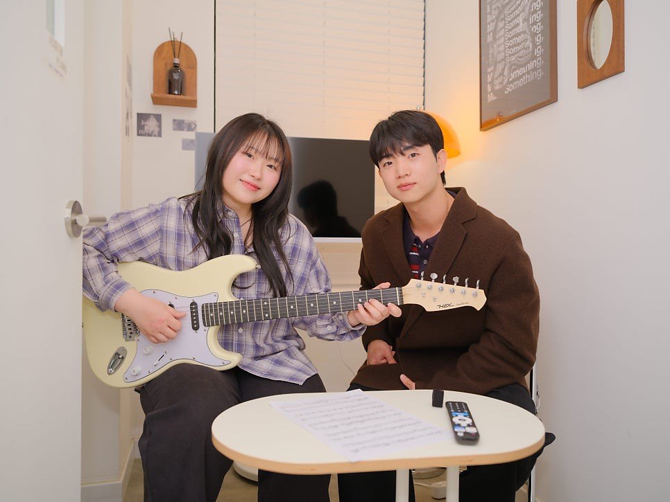흰 벽의 작은 레슨실에서 크림색 일렉기타를 든 수강생과 갈색 재킷의 강사가 나란히 앉아 있고, 앞의 둥근 탁자에 악보와 리모컨이 놓인 모습