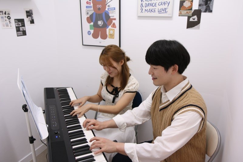 흰 벽 레슨실에서 니트 조끼 차림의 강사와 수강생이 나란히 앉아 디지털 피아노 건반을 치는 모습